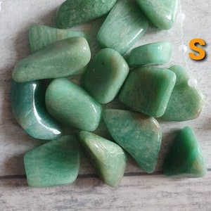 Può includere: Una collezione di pietre di avventurina verde lucidata. Le pietre variano per forma e dimensione, con un aspetto liscio e arrotondato. Il colore varia dal verde chiaro allo scuro. Un filo di perline bianche è visibile sullo sfondo, e la lettera "S" è in alto a destra.