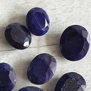 Peut inclure: Sept saphirs ovales et taillés, d'un bleu profond. Les pierres précieuses sont disposées sur une surface claire, chacune reflétant la lumière de manière unique. Les saphirs varient légèrement en taille et en clarté.