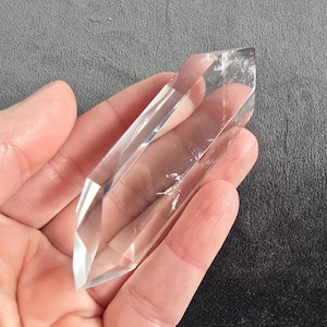 Könnte beinhalten: Ein klarer Quarz-Kristallpunkt mit mehreren Facetten. Der Kristall wird in einer Hand vor einem dunkelgrauen Hintergrund gehalten. Der Quarz ist transparent mit einigen inneren Einschlüssen.