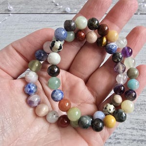 Könnte beinhalten: Ein mehrfarbiges Perlenarmband mit verschiedenen Steinperlen in Blau-, Grün-, Braun-, Gelb- und Lilatönen. Das Armband wird in einer Hand gehalten, wodurch die verschiedenen Farben und Texturen der Perlen zur Geltung kommen. Die Perlen haben einen Durchmesser von etwa 0,8 cm.