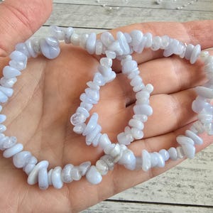 Puede incluir: Dos pulseras de virutas de piedras preciosas azul claro. Las pulseras están hechas de pequeñas piedras de forma irregular ensartadas. Las piedras tienen un color azul pálido y un aspecto ligeramente translúcido. Las pulseras se muestran en una mano.