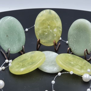 Könnte beinhalten: Eine Sammlung ovaler Steine in Grün- und Gelbtönen, präsentiert auf kleinen Metallständern. Einige Steine stehen aufrecht, andere liegen flach. Eine Schnur aus weißen Perlen schmückt die Szene vor dunklem Hintergrund.