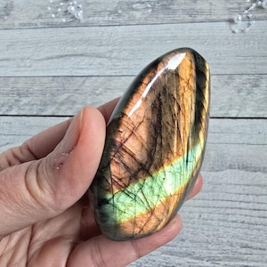 Può includere: Una pietra preziosa di labradorite lucidata, di forma ovale. La pietra mostra una miscela di colori, tra cui marrone, arancione, verde e blu, con una finitura lucida. La gemma è tenuta in una mano.