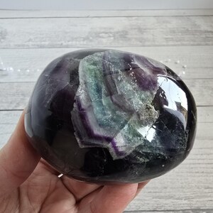 Pode incluir: Um cristal de fluorite polido, em forma oval. A pedra apresenta secções roxas escuras, roxas claras, verdes e transparentes. A superfície é lisa e reflectora, com variações naturais de cor e textura.