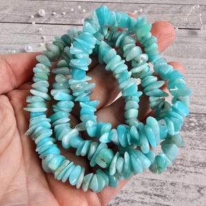 Peut inclure: Un fil de perles en copeaux d'amazonite bleu clair. Les perles sont un mélange de bleu clair et moyen, de forme légèrement irrégulière. Les perles sont enfilées ensemble, formant un collier ou un bracelet. Le fond est une surface en bois clair.