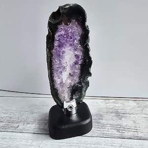 Könnte beinhalten: Eine polierte Amethyst-Geodenscheibe mit einem leuchtend violetten Kristallzentrum und einem dunklen, kontrastierenden äußeren Rand. Die Geode ist auf einem schwarzen, rechteckigen Sockel montiert und bildet ein auffälliges Ausstellungsstück. Natürlicher Dekostein.