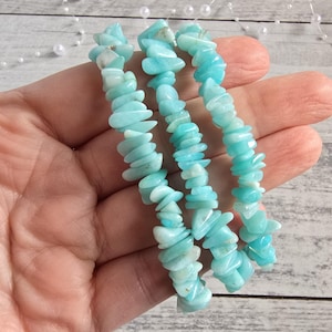 Op de afbeelding: Drie strengen lichtblauwe Amazoniet chip kralenarmbanden. De armbanden worden in een hand gehouden en tonen de onregelmatige, gepolijste steensplinters. De kralen hebben een mix van lichte en middelblauwe tinten, waardoor een getextureerde, natuurlijke uitstraling ontstaat.