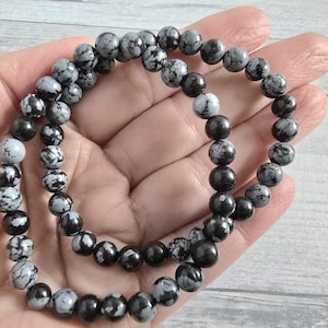 Könnte beinhalten: Zwei Armbänder aus Schneeflocken-Obsidian-Perlen. Die runden Perlen sind schwarz, grau und weiß. Die Armbänder werden in einer Hand gehalten.