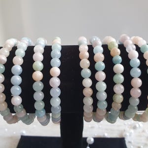 Könnte beinhalten: Mehrere Armbänder mit Perlen in Pastellfarben wie Rosa, Blau, Grün und Creme. Die Armbänder werden auf einem schwarzen Ständer präsentiert, der die Vielfalt der Farben und Texturen hervorhebt. Die Perlen scheinen Halbedelsteine zu sein.