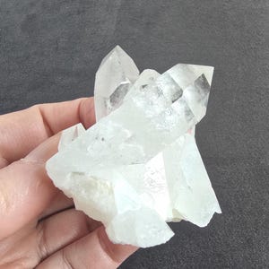 May include: A cluster of clear quartz crystals with sharp, pointed terminations. The crystals are translucent and appear to be held in a hand, showcasing their natural, raw form. The quartz has a slightly milky appearance, with some minor inclusions.
