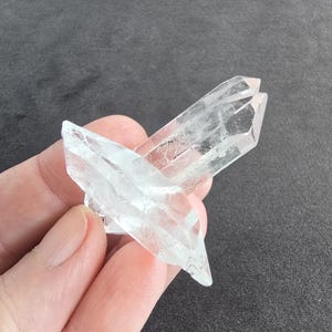 Könnte beinhalten: Ein Cluster aus klaren Quarzkristallen, gehalten in einer Hand vor einem dunkelgrauen Hintergrund. Die transparenten Kristalle haben mehrere Spitzen und natürliche Einschlüsse. Der Quarz reflektiert das Licht.