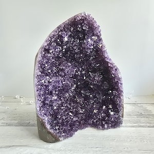 Könnte beinhalten: Eine große Amethyst-Geode mit einem tiefvioletten kristallinen Inneren aus gezackten, facettierten Spitzen. Die Außenseite des Steins ist eine glatte, dunkelgraue Gesteinsformation, die einen natürlichen Kontrast zwischen den rauen, funkelnden violetten Kristallen und der soliden, erdigen Basis des Mineralexemplars zeigt.