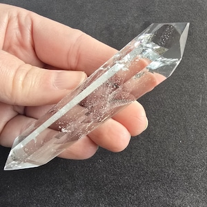 Op de afbeelding: Een heldere kwartskristalpunt met een facetgeslepen punt. Het kristal wordt in een hand gehouden tegen een donkere achtergrond. Het kwarts heeft interne insluitsels en een witte streep in het midden.