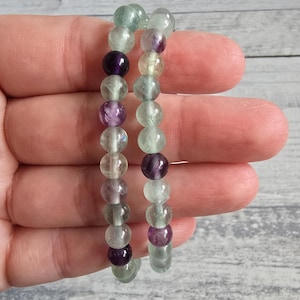 May include: Two beaded bracelets featuring a mix of purple, green, and clear fluorite beads. The bracelets are held in a hand, highlighting the colours and textures of the gemstones. The beads are approximately 0.8 cm in diameter.