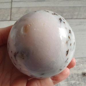 May include: A polished, spherical gemstone with a pale pink and white base colour, speckled with dark brown and black inclusions. The sphere is held in a hand, showcasing its smooth, reflective surface. The gemstone's size is approximately 7.6 cm in diameter.