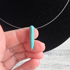 Può includere: Una collana con pendente turchese con una pietra sottile e allungata. La pietra ha un motivo marmorizzato ed è sospesa a un filo sottile color argento. La collana è esposta su uno sfondo nero.