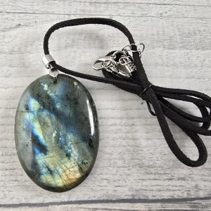 Peut inclure: Pendentif ovale en labradorite avec un cordon noir. La pierre précieuse affiche des teintes irisées bleues, vertes et jaunes. Le cordon est noué et possède un fermoir argenté.