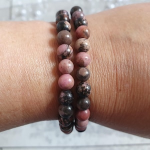Può includere: Due braccialetti di perline con perline sferiche rosa, marroni e nere. I braccialetti sono indossati su un polso, mostrando le varie sfumature e motivi delle perline di pietra naturale. Le perline hanno una finitura lucida.