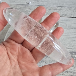 Può includere: Una bacchetta di cristallo di quarzo trasparente, allungata e liscia, tenuta in mano. Il cristallo presenta venature interne e una superficie lucida. Lo sfondo è una superficie di legno chiaro con una fila di perle.