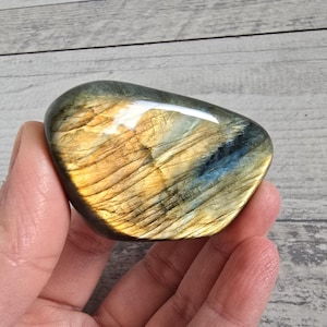 Può includere: Una pietra di labradorite lucidata, di forma ovale. La pietra mostra un'iridescenza scintillante con sfumature dorate, blu e verdi. La superficie è liscia e lucida. La pietra è tenuta in una mano.
