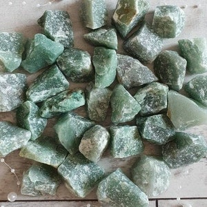 Op de afbeelding: Een verzameling ruwe, ongeslepen groene aventurijnstenen. De stenen variëren in grootte en vorm en vertonen een reeks lichte tot middelgroene tinten. Het natuurlijke, ongepolijste uiterlijk van de kristallen is duidelijk zichtbaar.