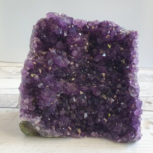May include: A large amethyst geode, displaying a rich purple colour. The crystal structure is dense and sparkling, with many individual points. The base of the geode has a green-grey hue. The overall shape is irregular and textured.