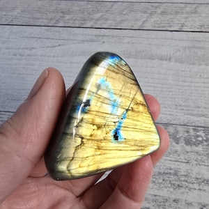 Può includere: Una pietra preziosa di labradorite lucidata, di forma triangolare. La pietra presenta una miscela di colori, tra cui giallo, grigio e blu, con riflessi iridescenti. La superficie ha una finitura liscia e lucida.
