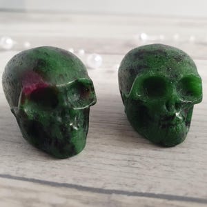 Op de afbeelding: Twee gebeeldhouwde edelsteen schedels. De schedels zijn diepgroen met rode en zwarte insluitsels. De schedels worden tentoongesteld op een lichtgekleurd houten oppervlak. Decoratieve items staan op de achtergrond.