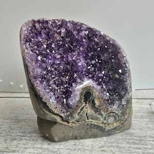 May include: A large amethyst geode with a polished, rounded form. The interior is filled with sparkling, deep purple crystals. The base is a rough, grey stone, set against a neutral background.