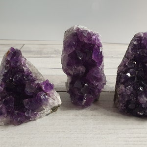 Könnte beinhalten: Drei Amethyst-Kristallcluster. Die Kristalle sind tiefviolett mit einer kristallinen Struktur. Die Cluster variieren in Größe und Form, mit einer rauen, natürlichen Textur. Der Hintergrund ist eine helle Holzmaserung.