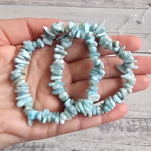 Könnte beinhalten: Zwei hellblaue Edelsteinarmbänder. Jedes Armband besteht aus kleinen, unregelmäßig geformten Steinen, die aneinandergereiht sind. Die Armbänder werden in einer Hand gehalten, um ihre Farbe und Textur zu zeigen. Die Steine haben eine Mischung aus hellblauen und weißen Farbtönen.