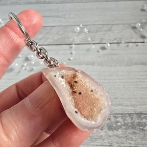 May include: A geode slice keychain. The geode is polished and semi-translucent, with a light pink exterior and a central cavity filled with tan and brown crystals. The keychain hardware is silver-coloured metal.