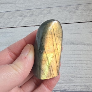 Può includere: Una pietra preziosa di labradorite lucidata di forma arrotondata. La pietra mostra un mix di colori, tra cui oro, verde e grigio, con linee interne visibili. La pietra preziosa è tenuta in una mano.