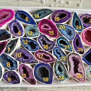 May include: A collection of colorful geode slices in a cardboard box. The geodes are numbered and feature a variety of colors, including pink, purple, blue, and teal. The geodes are displayed on a white base.