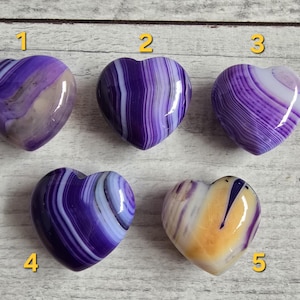Op de afbeelding: Vijf hartvormige stenen met verschillende patronen en kleuren. De stenen zijn voornamelijk paars, met witte, bruine en gele accenten. Elke steen heeft een uniek gestreept of gewerveld ontwerp. Ze liggen op een lichtgrijze houten ondergrond.