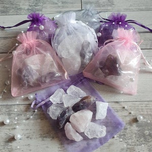 Könnte beinhalten: Eine Auswahl kleiner, durchsichtiger Beutel mit Kordelzug in Rosa, Lila und Weiß, gefüllt mit rohen Kristallen. Die Kristalle variieren in der Farbe, darunter Bergkristall, Amethyst und Rosenquarz. Perlenschnüre dienen als Dekoration.
