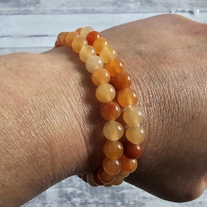 Può includere: Due braccialetti di perline arancioni e gialle sono indossati su un polso. Le perline rotonde sono in diverse tonalità di arancione, giallo e marrone. I braccialetti sono elastici e le perline sono infilate.