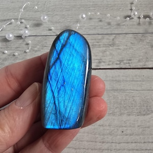 Könnte beinhalten: Ein polierter, blauer Labradorit-Edelstein mit schillernden Streifen. Der Stein wird in einer Hand gehalten, wodurch seine lebendigen, natürlichen Farben zur Geltung kommen. Der Edelstein hat eine glatte, abgerundete Form und ist ein schönes Beispiel für natürliche Kristallformationen.