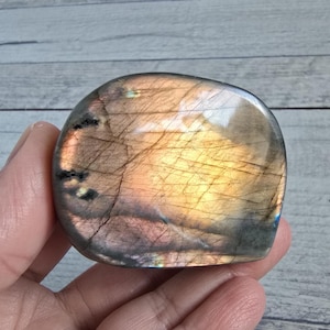 Può includere: Una pietra di labradorite lucidata di forma arrotondata. La pietra mostra un mix di colori oro, viola e grigio, con linee interne visibili. La superficie è liscia e riflettente, mettendo in risalto i motivi naturali della gemma.