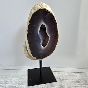 Puede incluir: Una rebanada pulida de geoda de ágata, que muestra un centro púrpura oscuro y un borde exterior beige claro. La geoda está montada sobre un soporte de metal negro, creando una pieza de exhibición llamativa. La piedra natural es un artículo decorativo único.