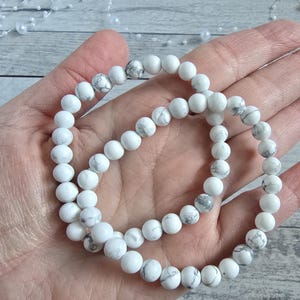 Può includere: Due braccialetti di perline bianche e grigie. I braccialetti sono fatti di perline rotonde e lucide. I braccialetti sono esposti su una mano, con una superficie di legno chiaro sullo sfondo.