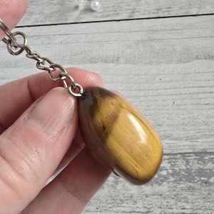 Op de afbeelding: Een gepolijste, langwerpige tijgeroog steen sleutelhanger. De steen heeft een warme, goudbruine kleur met donkerbruine strepen. De sleutelhanger hardware is van zilverkleurig metaal, inclusief een splitring en ketting.