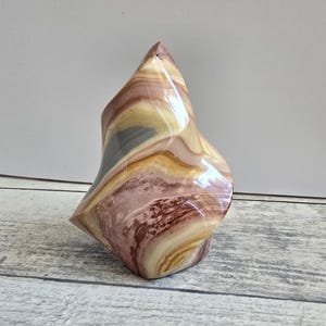 Può includere: Una scultura astratta lucidata nei toni del rosa, giallo e grigio. La pietra ha una forma liscia e fluida con una finitura lucida. La scultura è esposta su una superficie chiara con venature del legno.