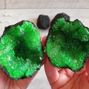 May include: Two halves of a geode with vibrant green crystal formations inside, held in hands. The outer edges are dark, contrasting with the bright green crystals. Two smaller, dark rocks are in the background.
