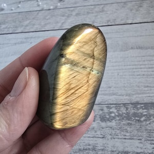 Può includere: Una pietra preziosa di labradorite lucidata, di forma ovale. La pietra mostra un mix di tonalità oro, grigio e blu con un effetto scintillante e iridescente. La superficie ha una finitura liscia e lucida.