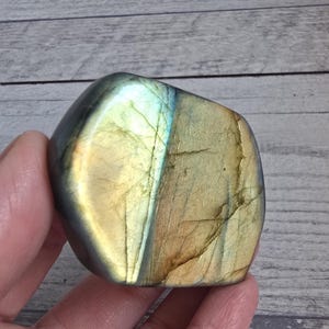 Può includere: Una pietra preziosa di labradorite lucidata con una forma liscia e arrotondata. La pietra mostra una miscela di colori, tra cui oro, blu e verde, con fratture interne visibili. La pietra preziosa è tenuta in mano, mostrando la sua bellezza naturale.