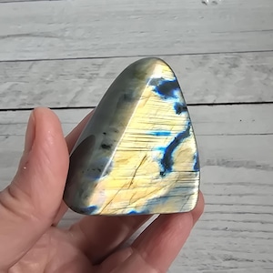 Può includere: Una pietra preziosa di labradorite lucidata, di forma triangolare, con una miscela di tonalità blu, gialle e grigie. La pietra è tenuta in mano, mostrando la sua superficie iridescente e i suoi motivi naturali. È un oggetto decorativo unico.