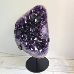 Könnte beinhalten: Eine große Amethyst-Geode mit tiefvioletten und schwarzen Kristallstrukturen. Die Geode ist auf einem schwarzen Metallständer mit rundem Fuß montiert, vor einem neutralen Hintergrund.
