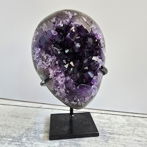 Könnte beinhalten: Eine große, polierte Amethyst-Geode, die ein tiefviolettes kristallines Inneres zeigt. Die Geode ist auf einem schwarzen Metallständer mit quadratischer Basis montiert. Das Äußere der Geode ist ein dunkles, gesprenkeltes Grau, das mit den leuchtenden violetten Kristallen kontrastiert.