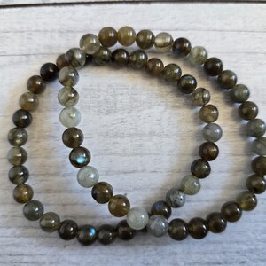 Könnte beinhalten: Zwei Armbänder aus runden, polierten Labradoritsteinen. Die Steine variieren in der Farbe von Dunkelbraun bis Hellgrau, mit blauen und grünen Reflexen. Die Armbänder werden auf einer hellen Holzoberfläche präsentiert.